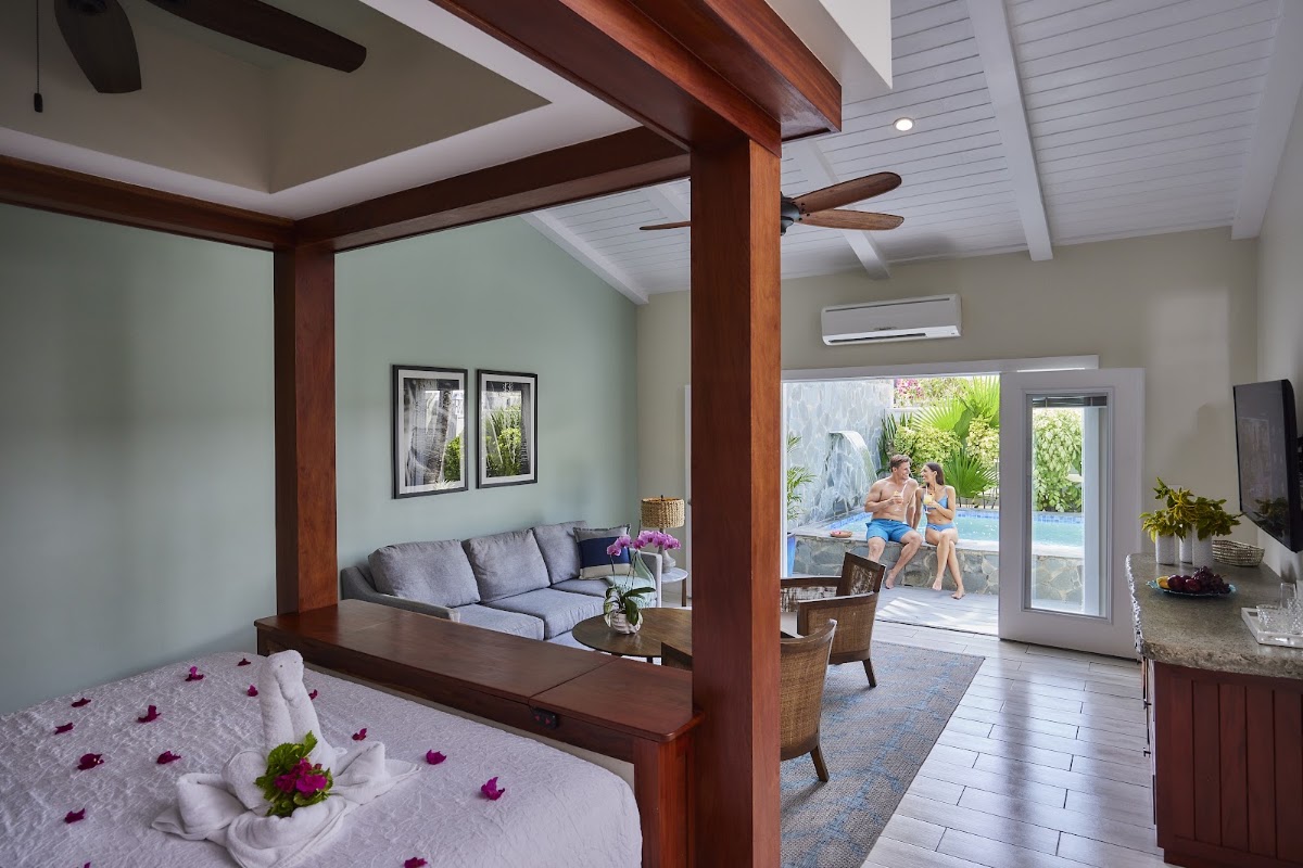 Spacious suite with four-poster bed and purple petals, opening to living area with sofa and glass doors overlooking pool and guests outside.