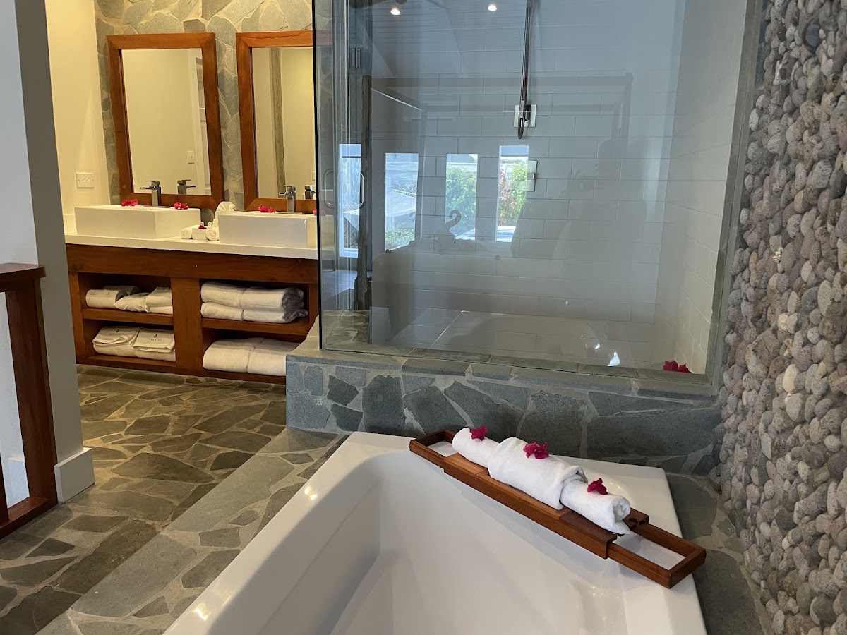 Bathroom with soaking tub in foreground, glass-enclosed shower, dual vanity with mirrors, and rolled towels on wooden tray at Serenity at Coconut Bay.