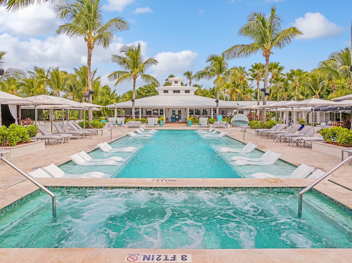 Turquoise pool with spa jets in foreground, main swimming pool beyond, flanked by lounge chairs and palm trees, with a white main building in the background at Serenity at Coconut Bay.