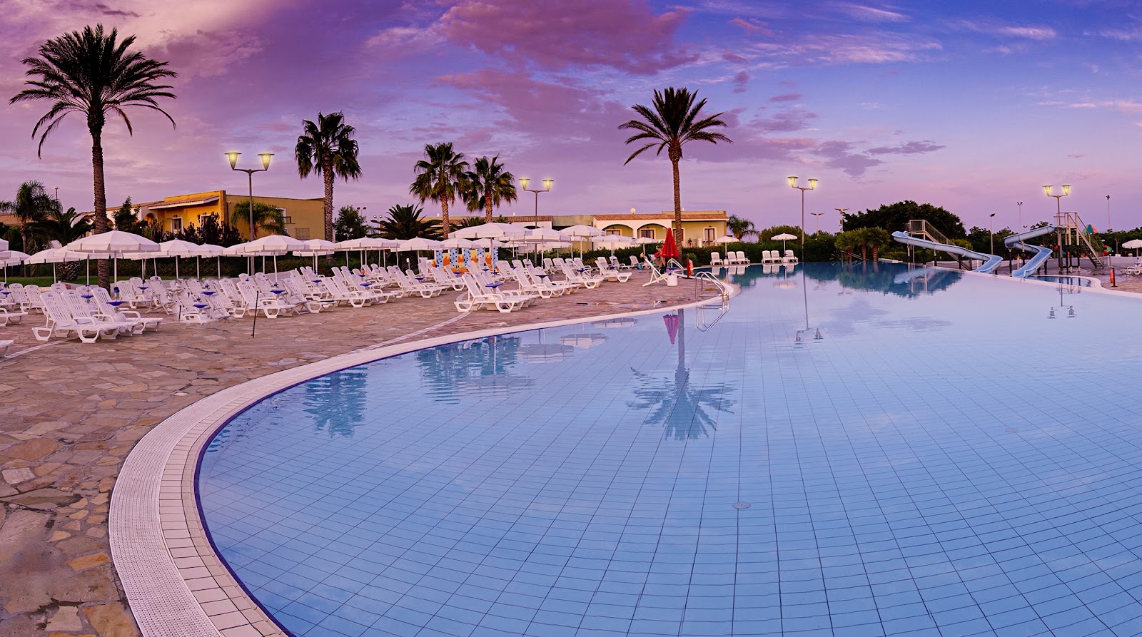 Blue pool and water slide at dusk with sandy beach, lounge chairs, and calm sea beyond at Serenusa Resort.