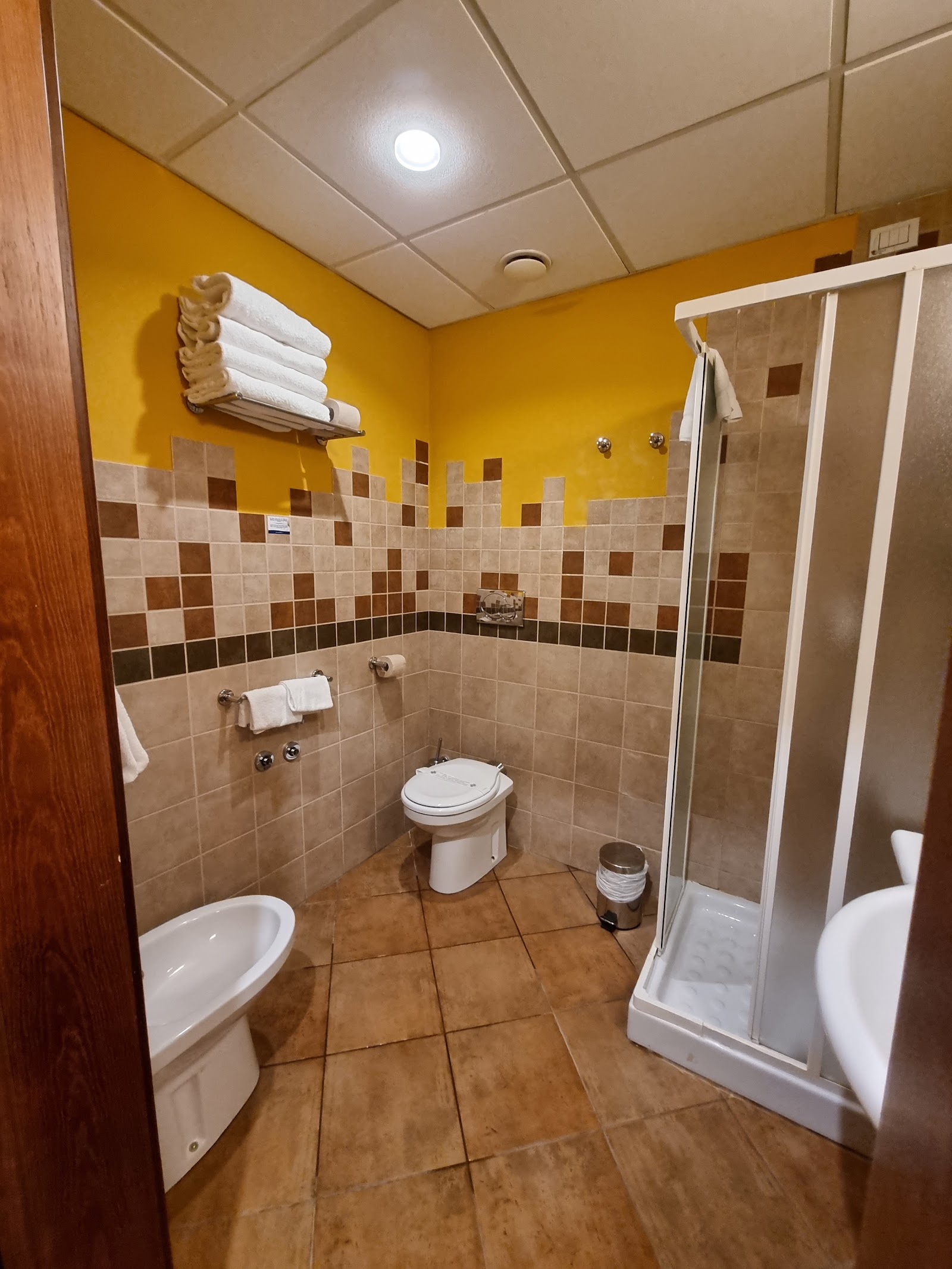 Bathroom with yellow upper walls and brown-and-tan tile, featuring a toilet, sink, shower stall, and white towels on a wall rack.