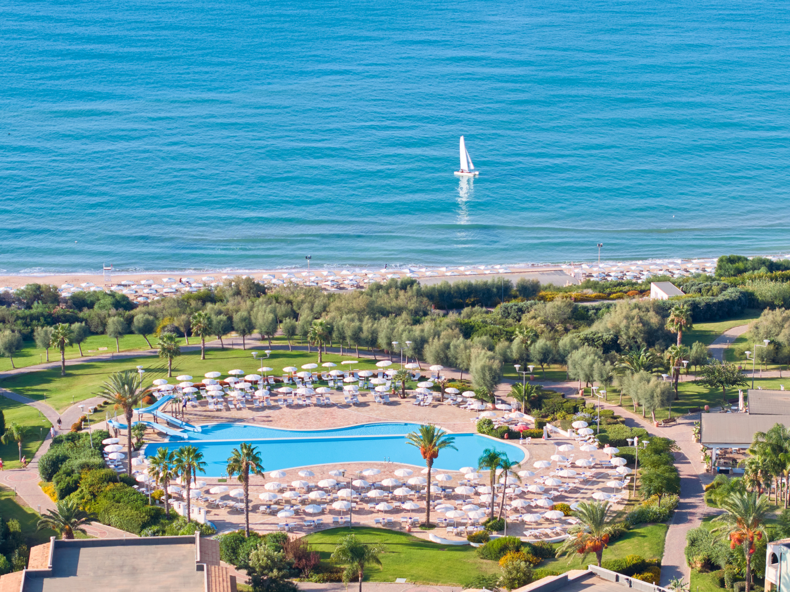 Aerial view of Serenusa Resort's pool complex with loungers and umbrellas, surrounding gardens, and beach beyond with sailboat on turquoise water.