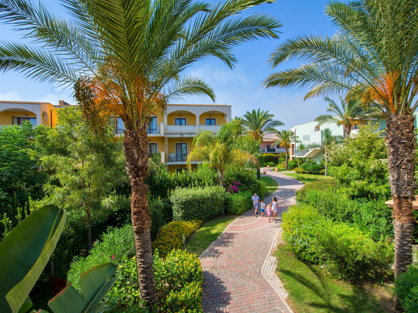 Brick pathways wind through lush landscaping with palm trees and flowering shrubs toward yellow and white resort buildings under clear blue sky.
