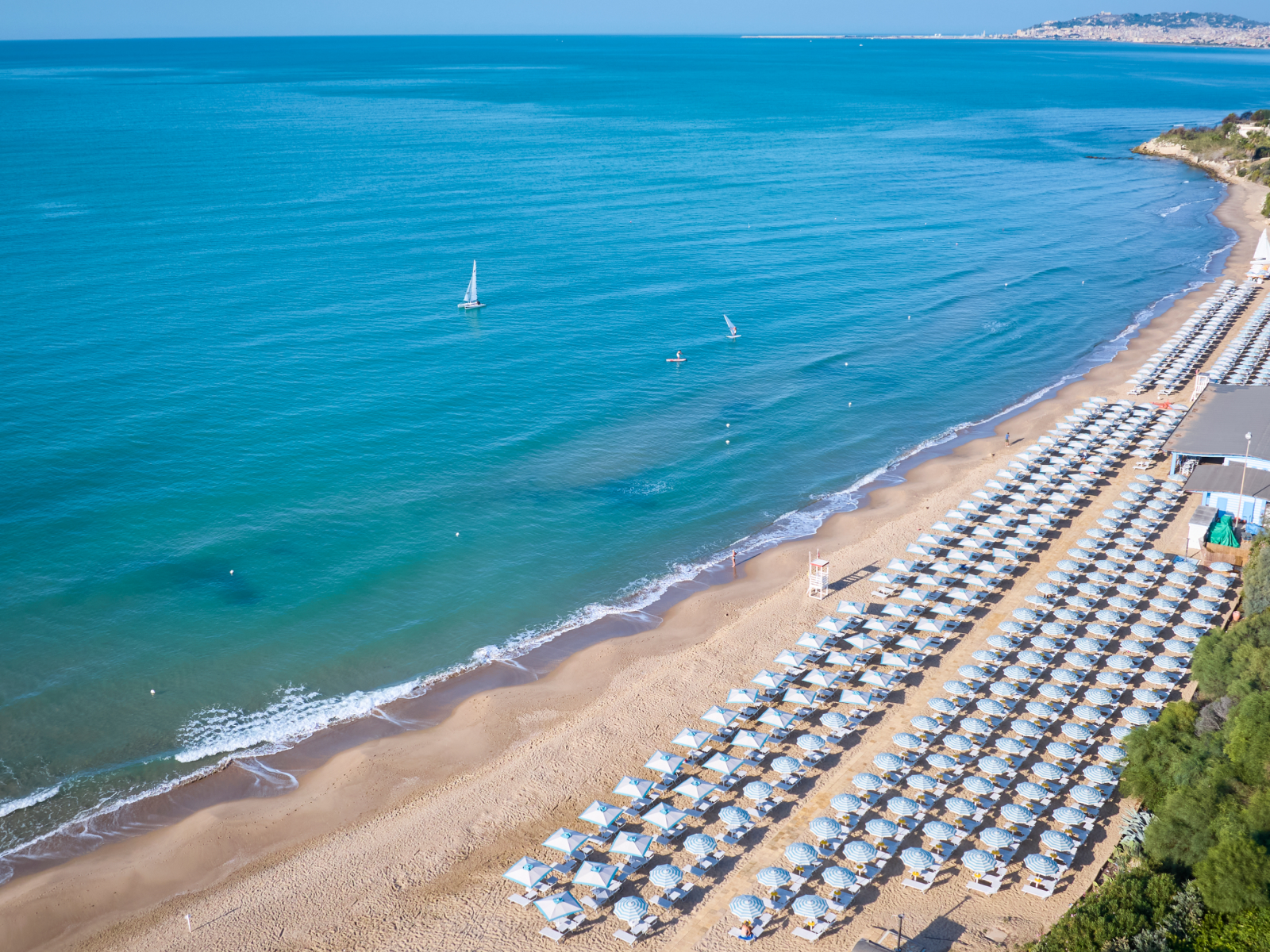 Sandy beach lined with blue and white cabanas at Serenusa Resort, with sailboats in turquoise water and coastal hills beyond.