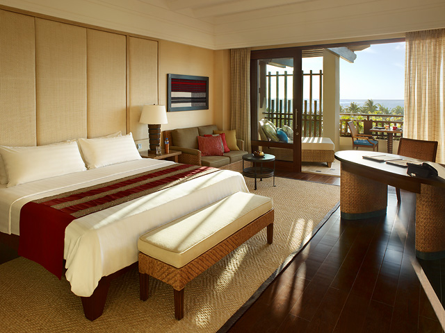 Spacious resort suite with king bed, cream walls, wooden furnishings, and ocean view from balcony overlooking palm trees and water.