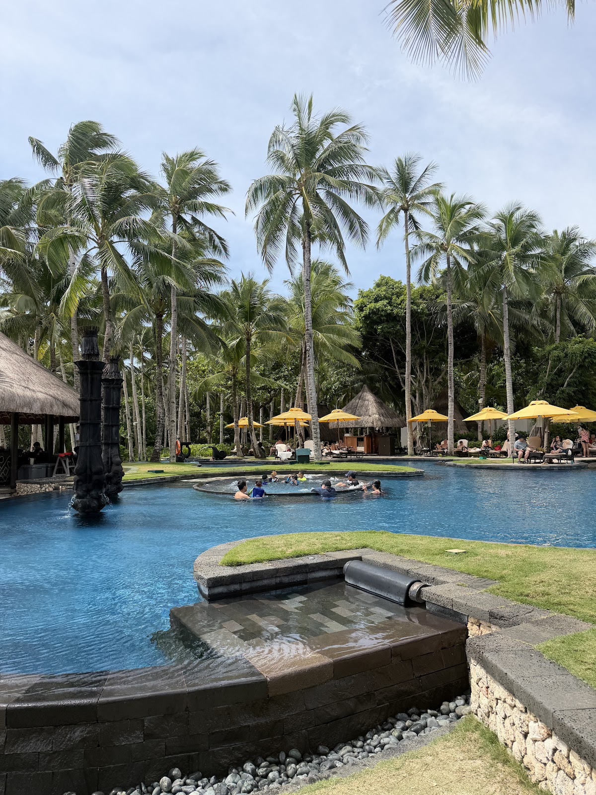Resort pool surrounded by tall coconut palms, with guests swimming and relaxing under yellow umbrellas on the lawn.