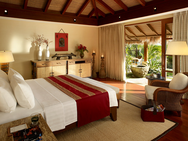 Spacious resort bedroom with wooden beamed ceiling, white bedding with red runner, and sliding glass doors opening to tropical garden views.