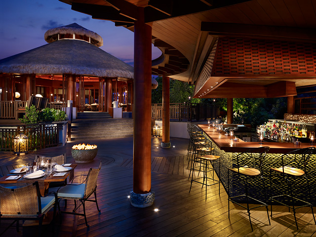 Twilight view of an outdoor dining area with thatched-roof restaurant, wooden deck, bar seating, and lit fire bowls at Shangri-La Boracay Resort & Spa.
