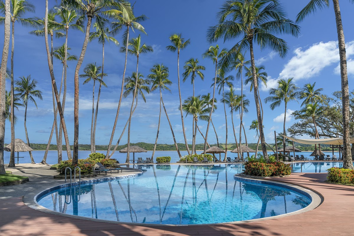 Curved pool with palm trees and lagoon view at Shangri-La Yanuca Island, with thatched structures and lounge chairs visible along the waterfront.