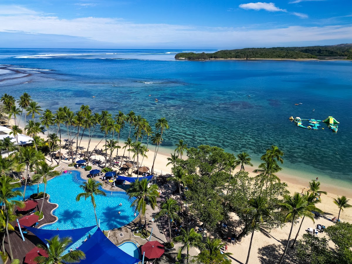 Resort pool and beach with palm trees overlooking turquoise ocean, forested island, and water sports activities.