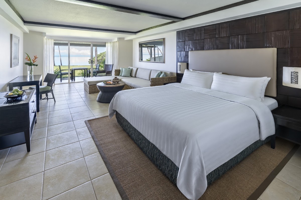 Resort suite with king bed, sitting area, and ocean view through glass doors opening to a balcony overlooking Fiji waters.