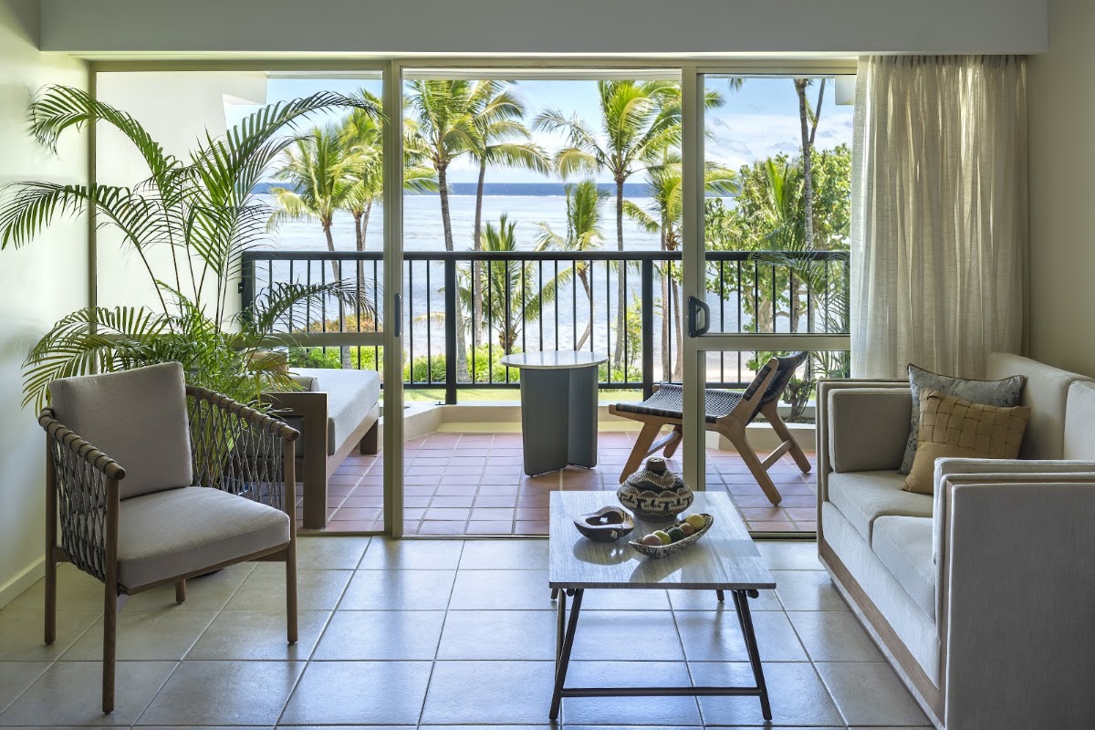 Resort suite interior with ocean view through glass doors, palm trees on balcony, beige furniture, and water visible beyond tropical vegetation.