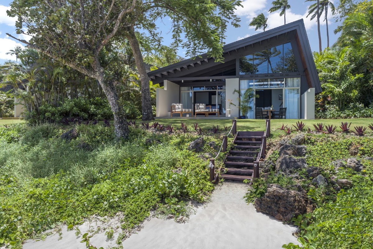 Modern beach villa with black roof and large windows surrounded by tropical vegetation, wooden steps leading through landscaped grounds with purple plants and palm trees.