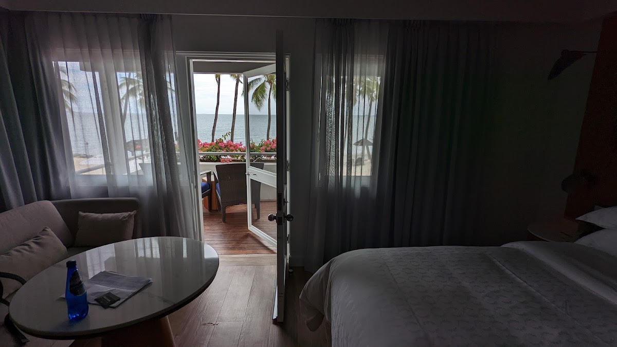 Resort room with bed and sitting area opening onto a balcony with ocean view, palm trees, and red flowers.