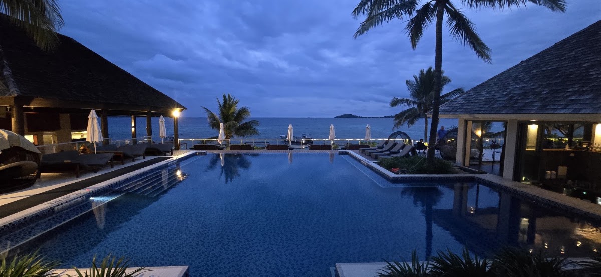 Illuminated infinity pool at dusk overlooking the ocean, flanked by palm trees, lounge chairs, and thatched-roof pavilions at Sheraton Fiji Golf & Beach Resort.