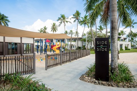 Children's playground with yellow slide at Sheraton Fiji Golf & Beach Resort, surrounded by palm trees and a wooden fence.