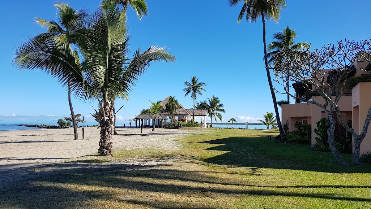 Palm trees and manicured lawn slope toward a sandy beach and thatched-roof pavilion at Sheraton Fiji Golf & Beach Resort, with ocean and sky beyond.