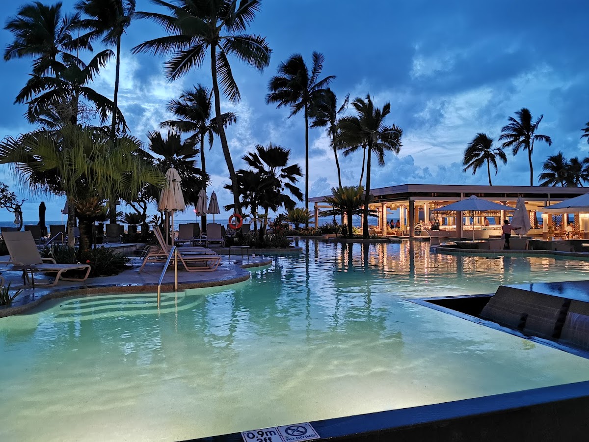 Sheraton Fiji Golf & Beach Resort's lagoon pool glows turquoise at dusk, surrounded by tall palms, lounge chairs, and an illuminated beachfront restaurant.
