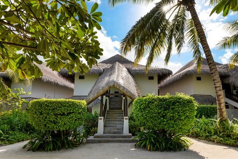 Thatched-roof bungalows with white walls frame a central entrance at Sheraton Maldives Full Moon Resort, surrounded by palm trees and manicured hedges.