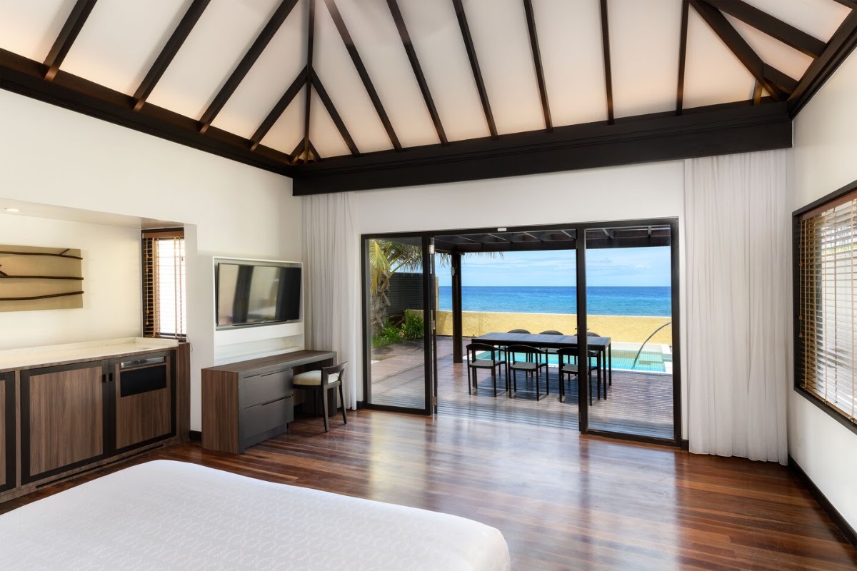 Modern beach villa interior at Sheraton Maldives Full Moon Resort with wooden floors, skylight ceiling, and ocean view through glass doors.