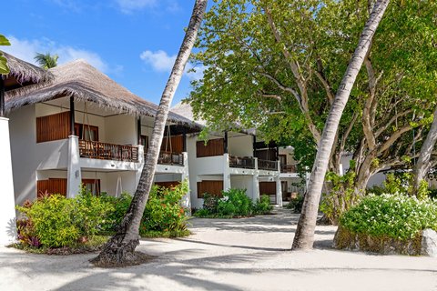 White bungalows with thatched roofs line a sandy pathway shaded by tall palm trees at Sheraton Maldives Full Moon Resort & Spa.