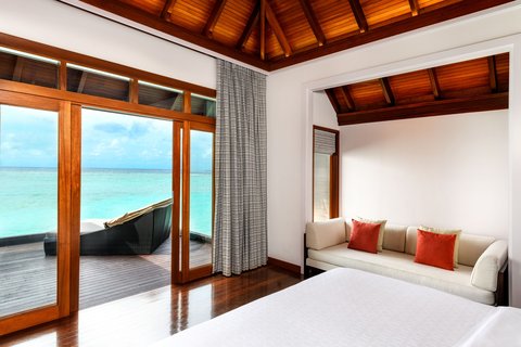 Overwater bungalow bedroom at Sheraton Maldives Full Moon Resort & Spa with wooden ceiling, bed, and glass doors opening to turquoise lagoon and wooden deck.
