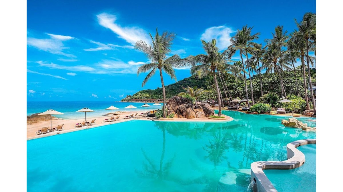 Turquoise pool with curved loungers and palm trees frames a sandy beach with umbrellas and ocean view at Sheraton Samui Resort.
