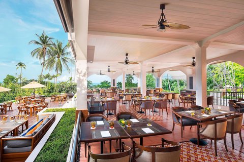Sheraton Samui Resort stretches along a sandy beach with loungers, backed by palm trees and jungle-covered hills under blue sky.
