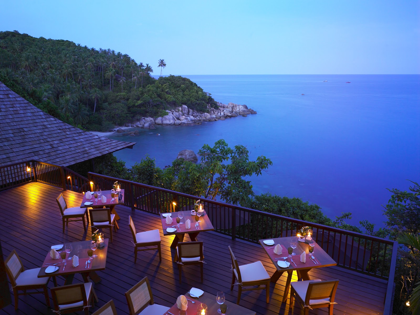 Candlelit dining tables on a wooden deck overlooking a rocky cove and deep blue ocean at dusk.