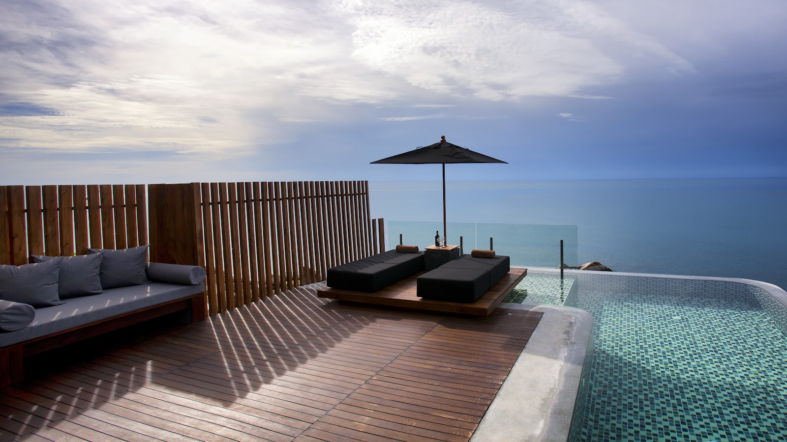 Curved infinity pool with turquoise water overlooks a calm tropical sea at Silavadee Pool Spa Resort, with a dark-roofed bungalow and wooden deck visible.