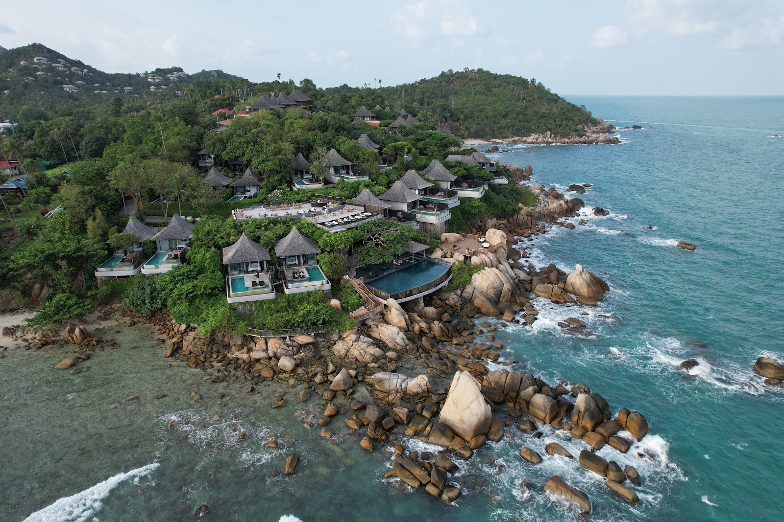 Silavadee Pool Spa Resort nestled on a forested hillside with thatched-roof villas overlooking turquoise waters and large granite boulders along the rocky coastline.