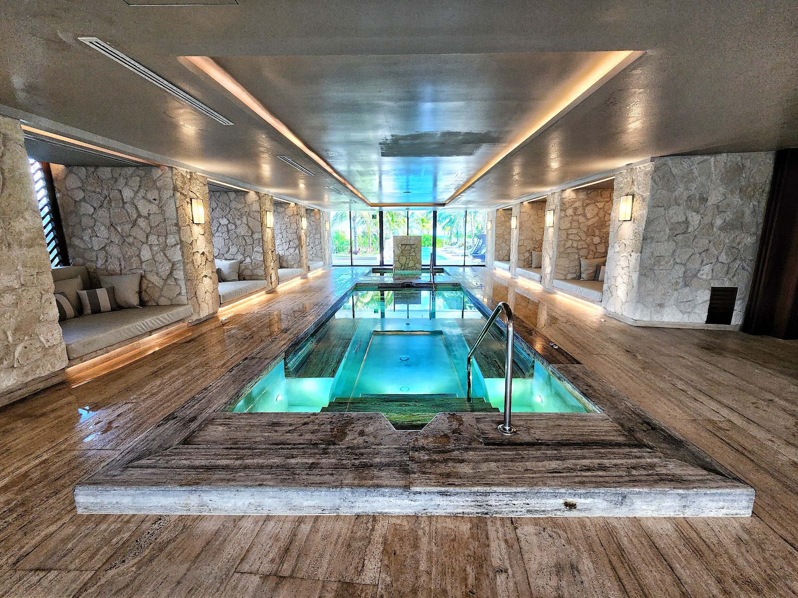 Indoor swimming pool with turquoise water, stone walls, wooden flooring, built-in seating alcoves, and glass doors opening to the grounds at SLS Playa Mujeres.