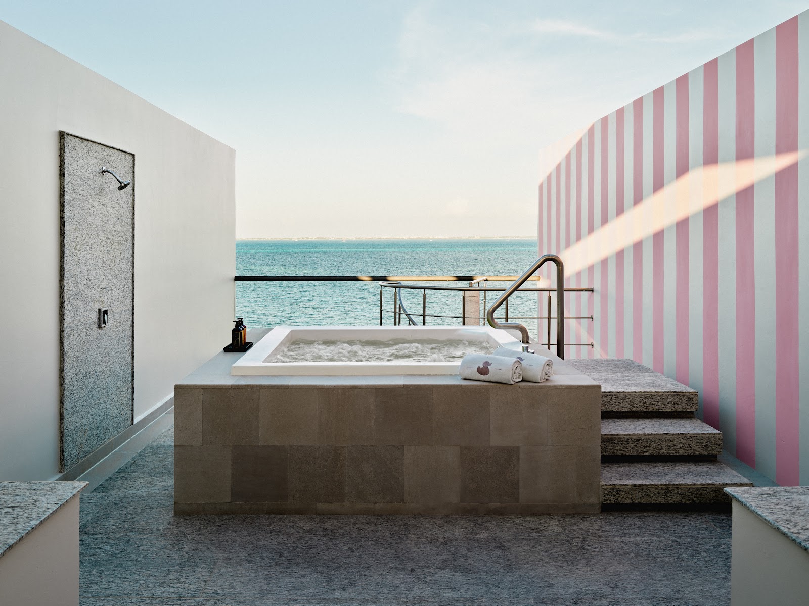 Outdoor soaking tub with ocean view at SLS Playa Mujeres, featuring pink and white striped walls and towels nearby.