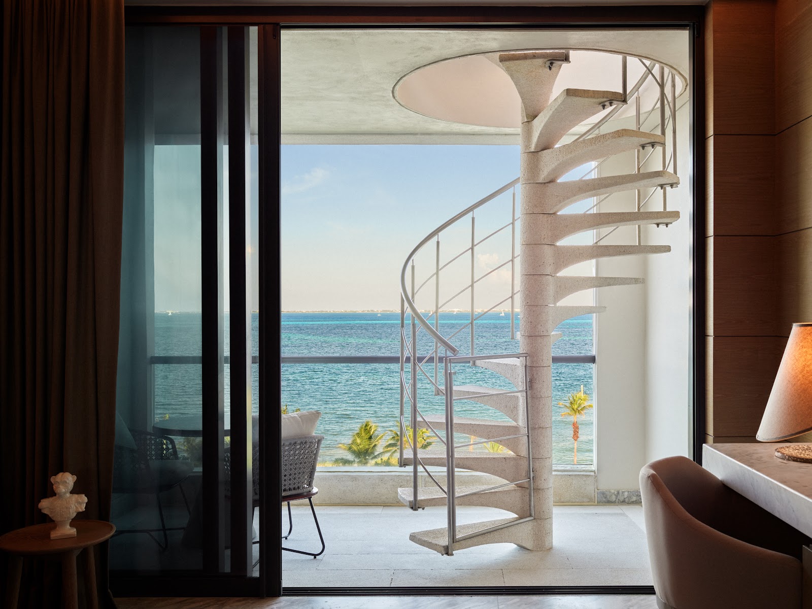 Resort room with open doors framing a spiral staircase, balcony, and turquoise Caribbean Sea beyond.