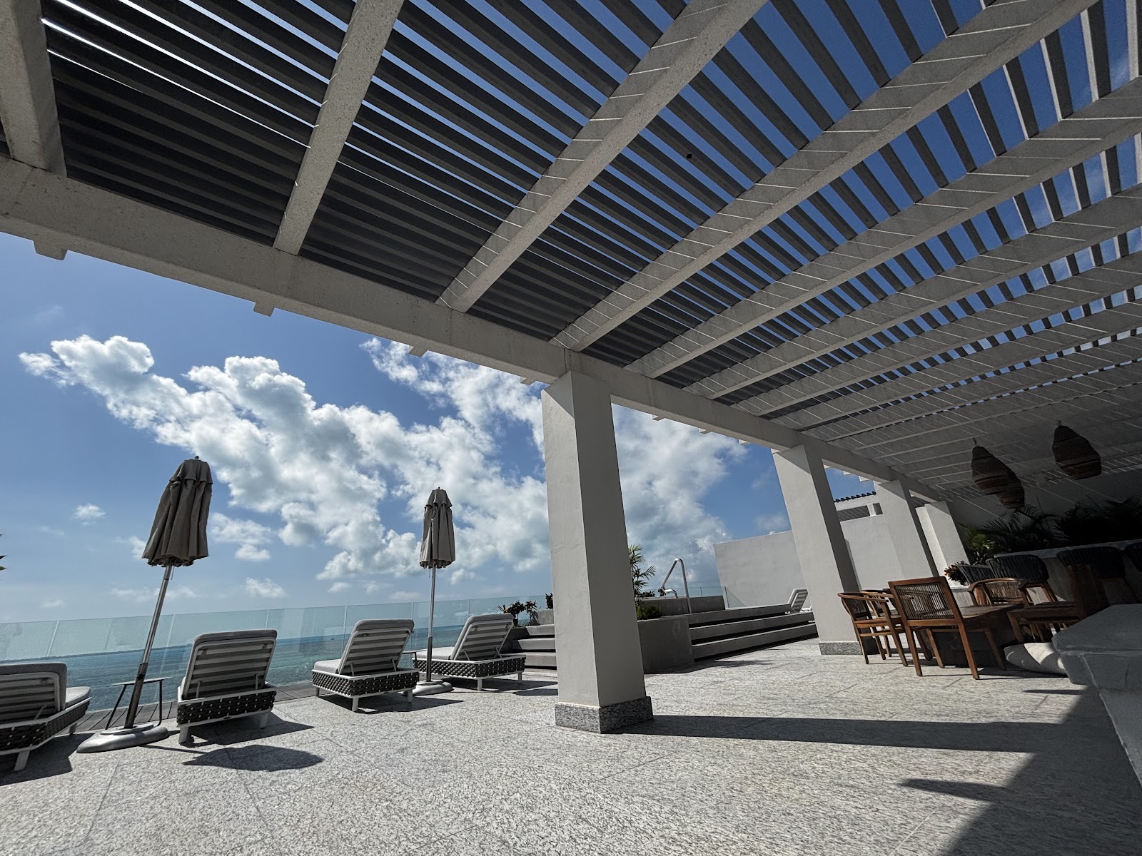 Shaded oceanfront terrace at SLS Playa Mujeres with lounge chairs, closed umbrellas, and wooden dining furniture beneath a slatted pergola.