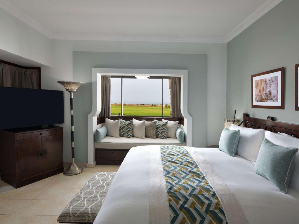 Guest room with queen bed, window seat overlooking green fields, TV, and sage green walls at Sofitel Agadir Royal Bay Resort.