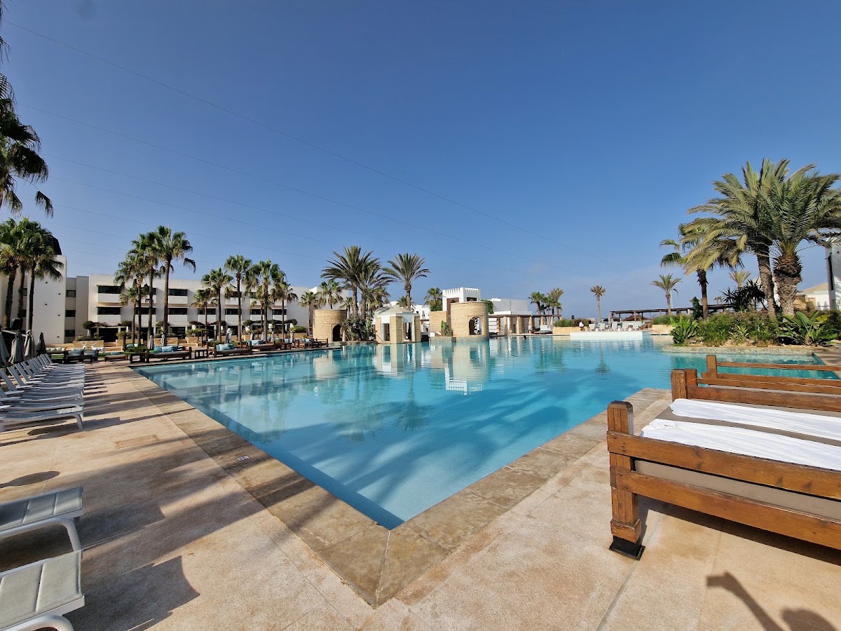 Large rectangular pool with turquoise water surrounded by loungers and wooden benches, with palm trees and beige resort buildings under clear blue sky.