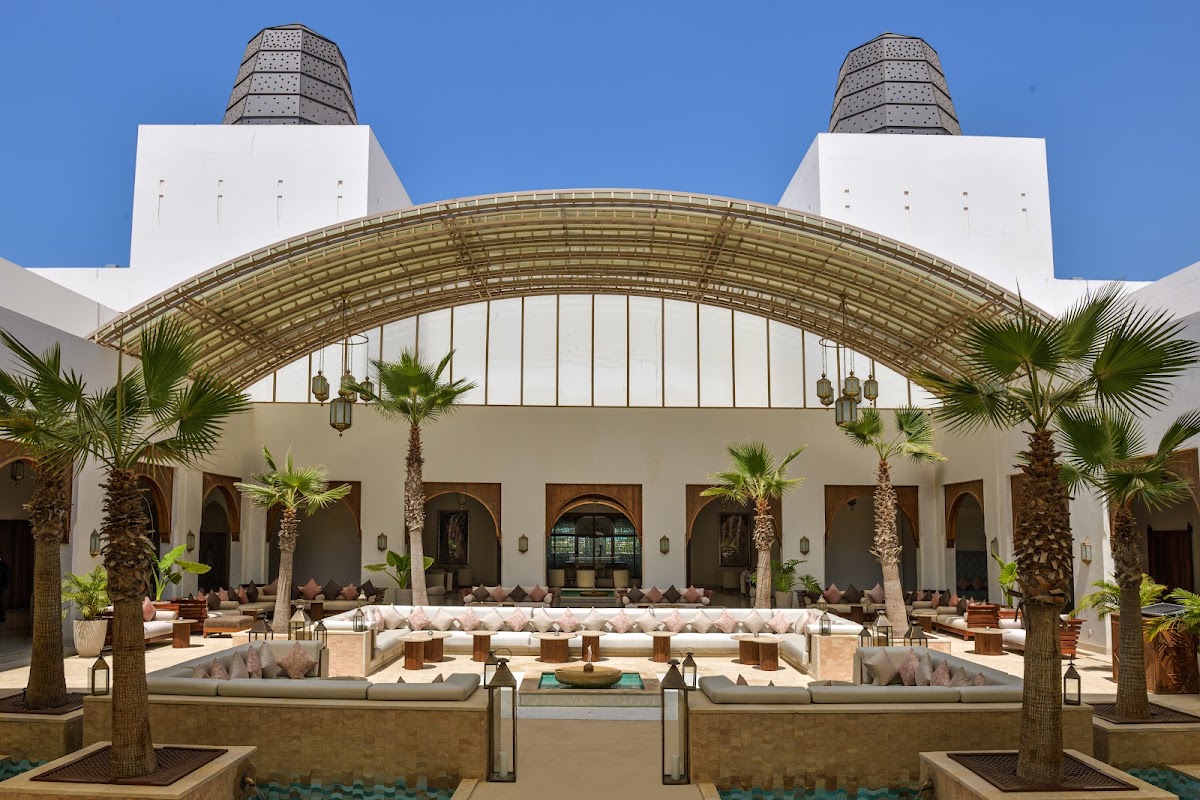 Sofitel Agadir Royal Bay Resort courtyard with white arched building, two towers, palm trees, sunken seating areas, and hanging lanterns under blue sky.