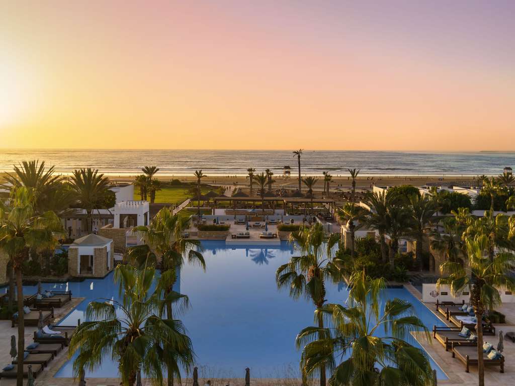 Large lagoon-style pool surrounded by palm trees and loungers at Sofitel Agadir Royal Bay Resort, with beach and ocean beyond at sunset.