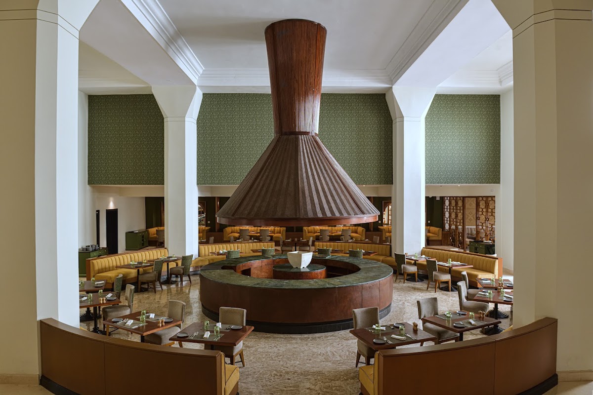 A circular dining room with a large conical wood hood suspended over a central fireplace, surrounded by mustard-yellow banquettes and tables set for service.