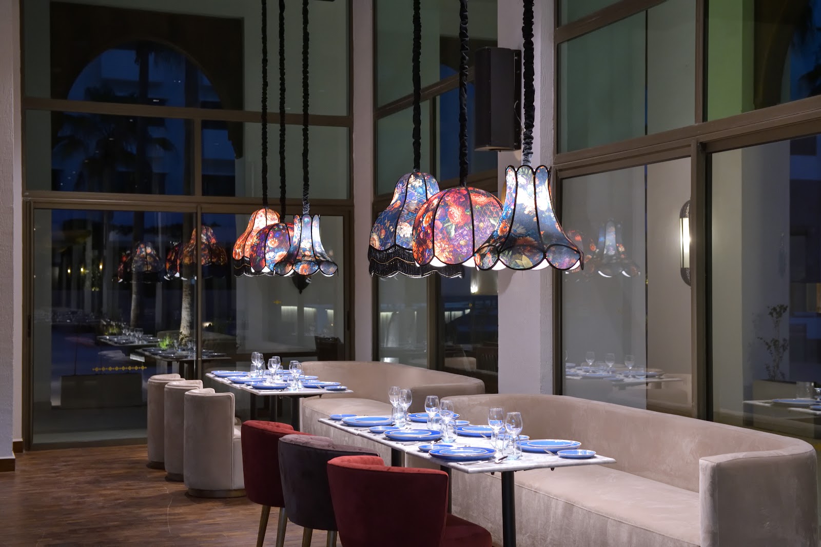 Modern dining room with cream banquettes, burgundy chairs, and colorful stained-glass pendant lamps hanging above a set table with blue napkins.