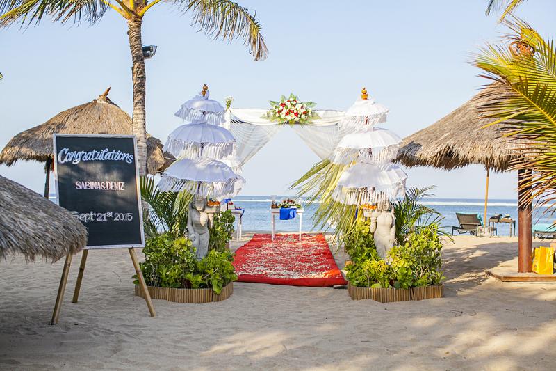 Beach wedding ceremony setup with white draped arch, red petal aisle, and thatched-roof palapas at Sol by Meliá Benoa Bali.