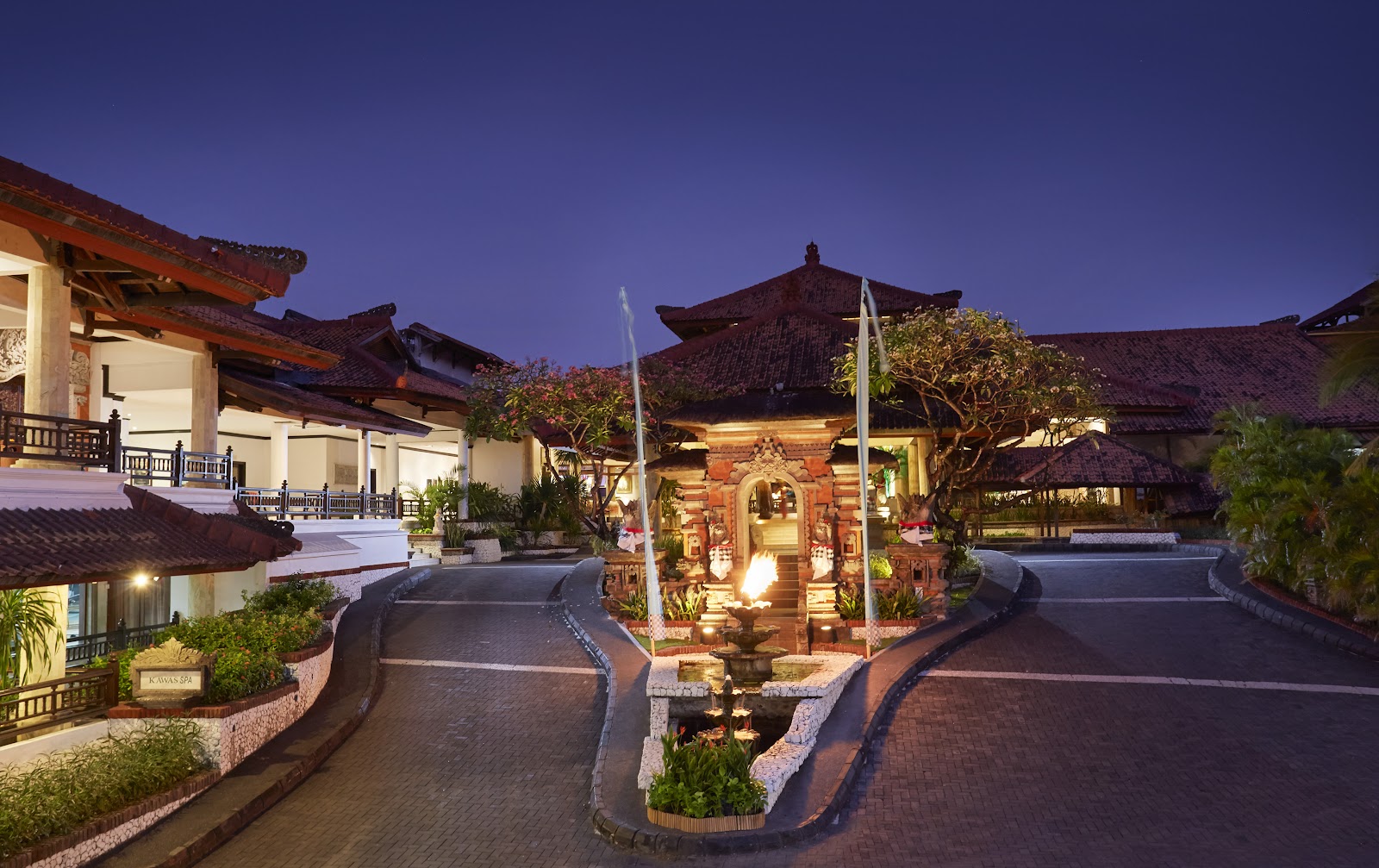 Balinese-style resort buildings with dark tile roofs line a curved brick pathway at dusk, with a lit stone gateway structure in the center and tropical plants throughout.