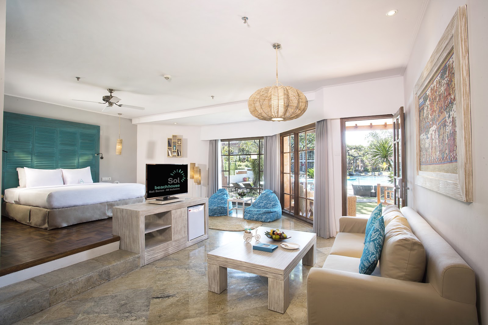 Spacious resort suite with king bed, teal headboard, woven pendant light, beige seating area, and glass doors opening to ocean views.