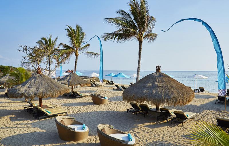 Beach at Sol by Meliá Benoa Bali with thatched-roof palapas, loungers, palm trees, and calm ocean beyond.