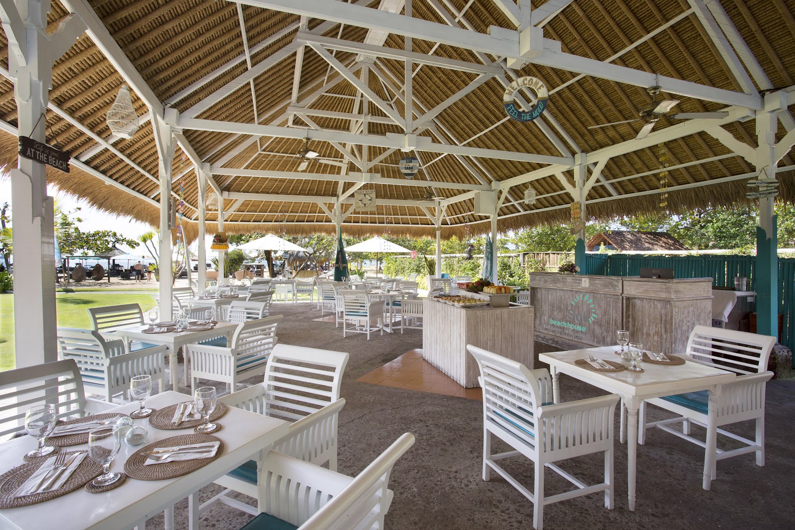 Large covered dining pavilion with white wooden beams, thatched roof panels, and set tables with white chairs overlooking manicured grounds at Sol by Meliá Benoa Bali.