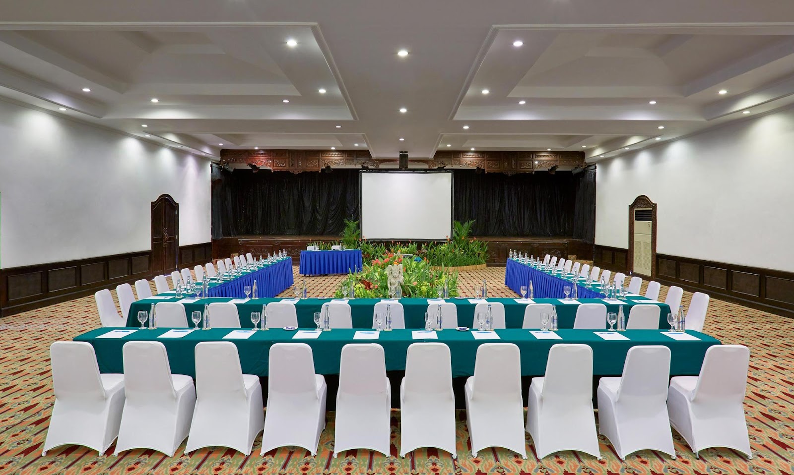 Banquet hall with long tables dressed in teal linens, white chair covers, projection screen, and tropical floral centerpieces.