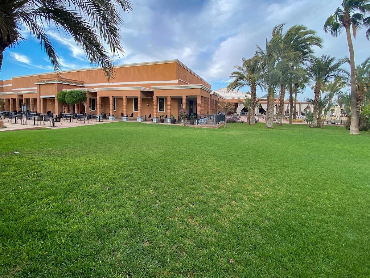 Large manicured lawn in front of terracotta building with palm trees at Sol Oasis Marrakech, outdoor seating visible under covered patio.