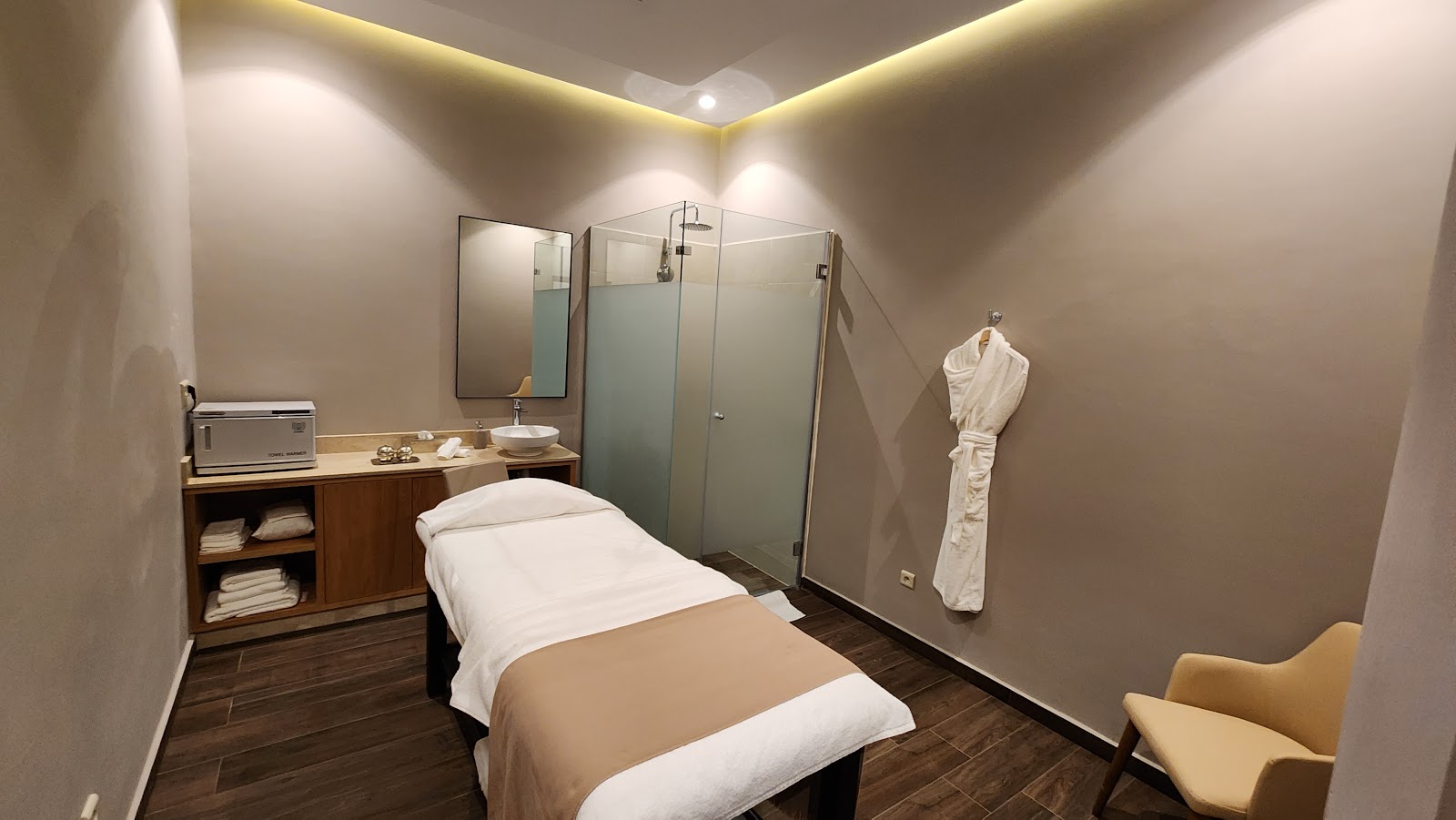 Spa treatment room with massage bed, glass shower enclosure, wooden vanity with toiletries, white robes, and ambient ceiling lighting.