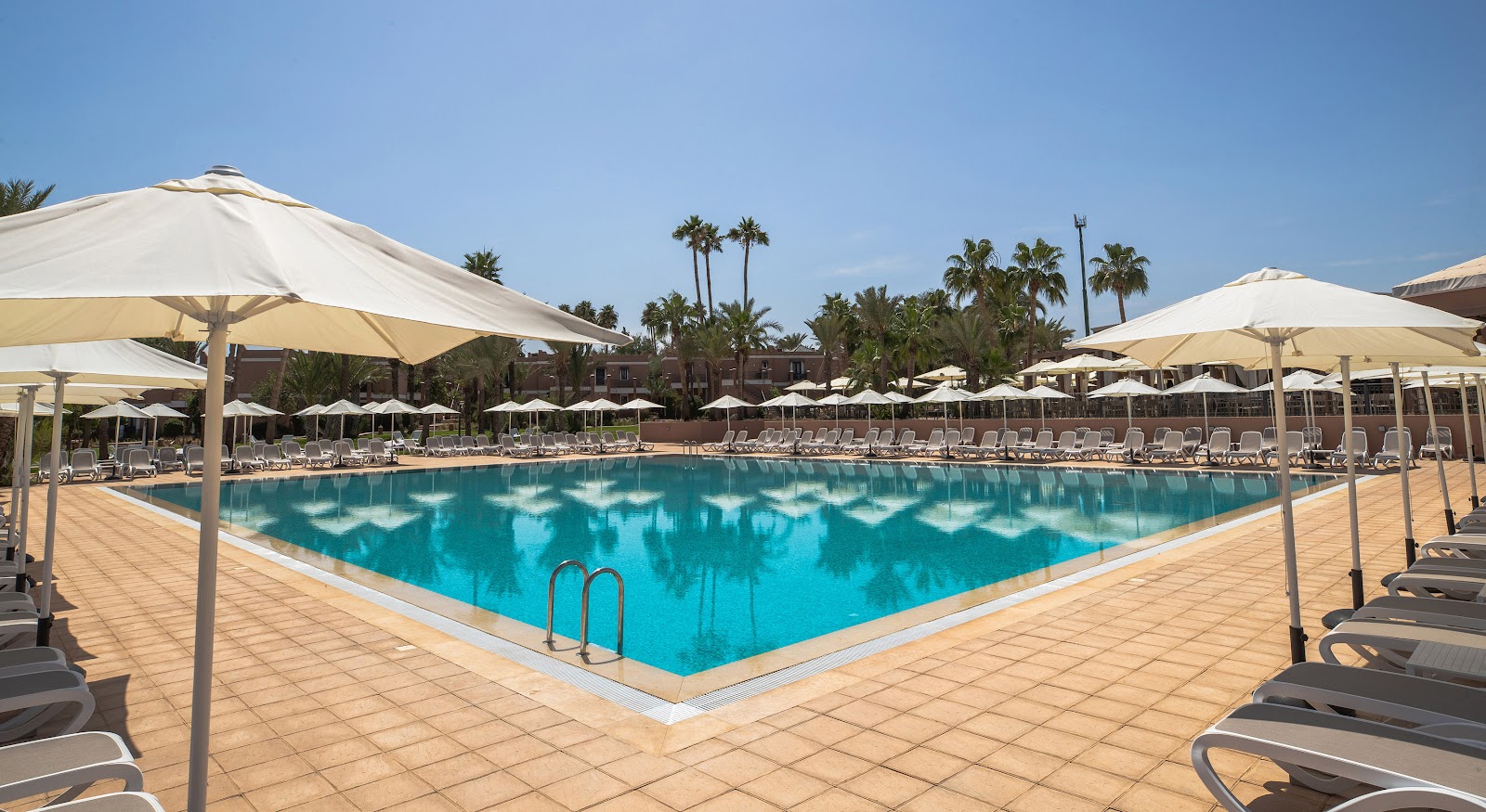 Large rectangular pool surrounded by tan umbrellas and loungers on terracotta tile deck, with palm trees and resort buildings in background under clear blue sky.